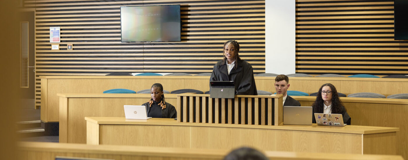 A student in a legal attire stands at a podium in a lecture hall, next to a desk. Seated at the desk are three students using laptops.
