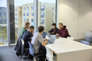 Five students sit around a table in the Learning Hub. they are working on laptops and are engaged in discussion. Behind them, there is a large floor-to-ceiling window that displays views of Waterside campus.