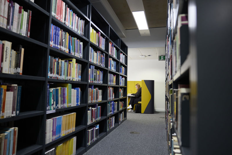 A student sits at a yellow study booth in the Learning Hub, surrounded by bookshelves filled with various books.