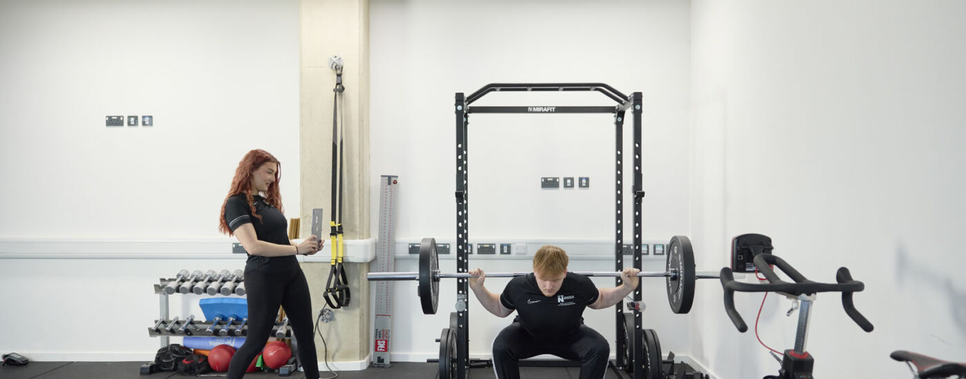 A person squats with a barbell across their shoulders in UON’s SIRC while another person stands nearby, observing. Exercise equipment including a bike, dumbbell rack, and squat rack are visible in the background.