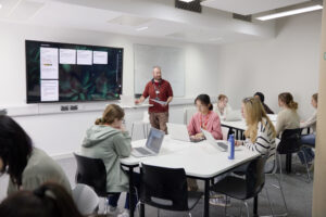 An Education classroom with students seated at tables with laptops, and an instructor standing at the front near a large screen displaying digital content.