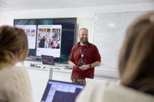 A smiling academic gives a presentation in a classroom. Two people sit with laptops. A large screen and whiteboard are in the background.
