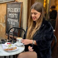 Yulianna Skakun wears a black coat and sits at a table with a cappuccino and a croissant topped with strawberries and powdered sugar in an outdoor cafe. A sign advertising taglieri misi is in the background.