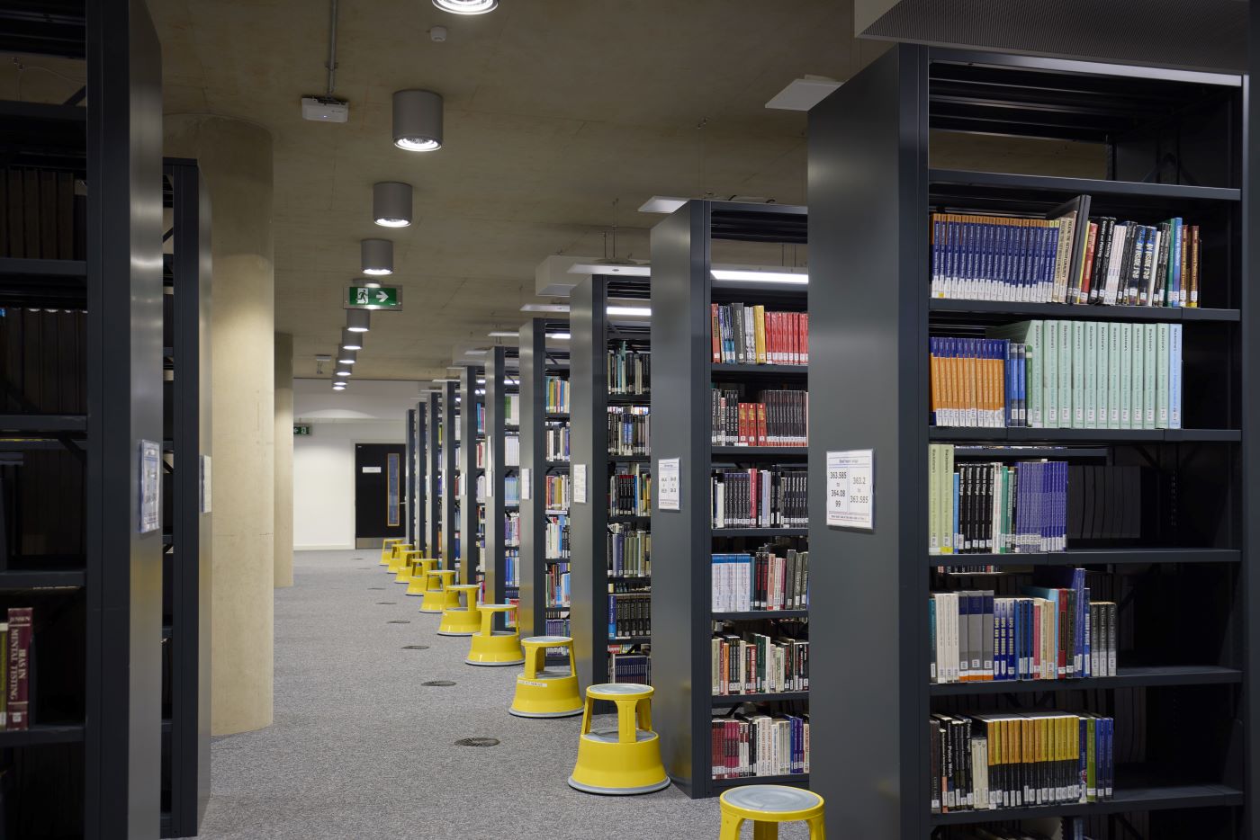 Library aisle with tall shelves filled with assorted books. Yellow stools are aligned along the floor. Ceiling lights illuminate the space.