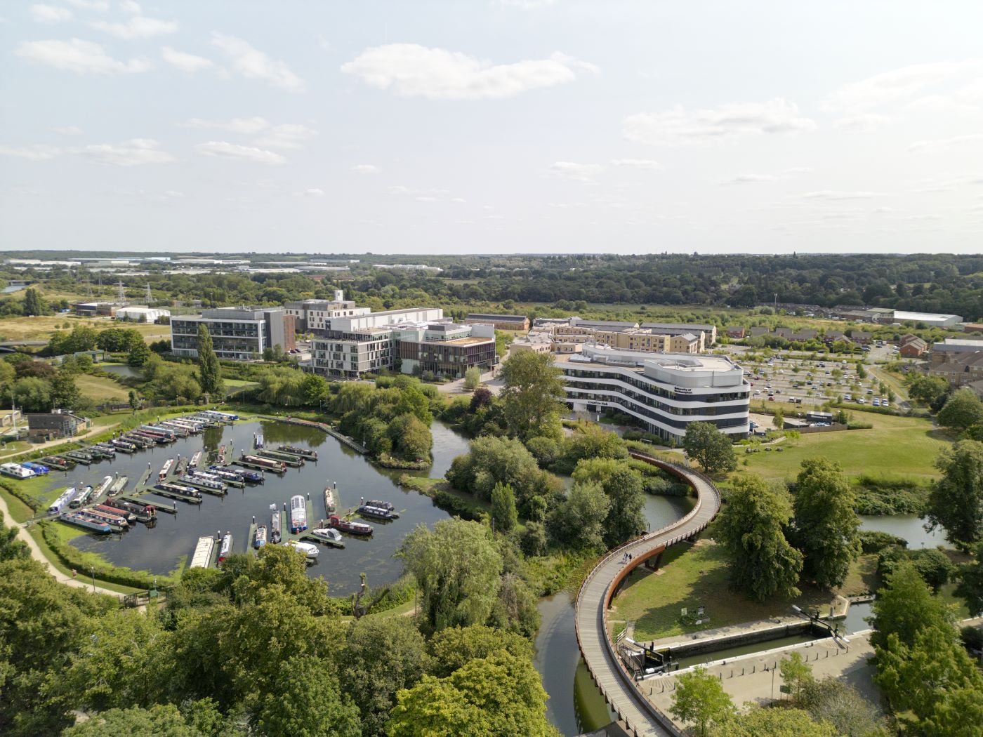 Aerial view of a Waterside campus with several buildings, a marina with boats, a curved footbridge, and surrounding greenery under a partly cloudy sky.