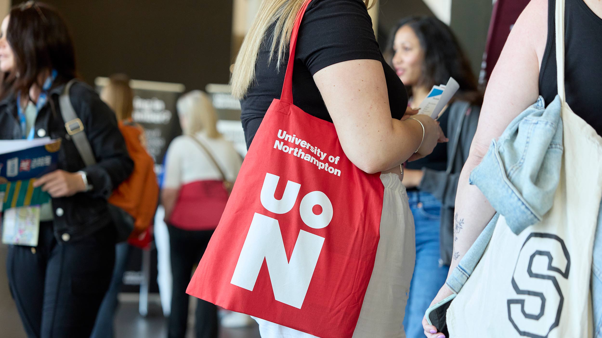 A visitor with a bright red University of Northampton tote bag holds an information leaflet at an indoor event; other individuals are blurred in the background.