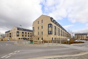 A modern, three-story apartment building with light brick exterior and dark-framed windows, situated on a corner with a paved road and some landscaping.