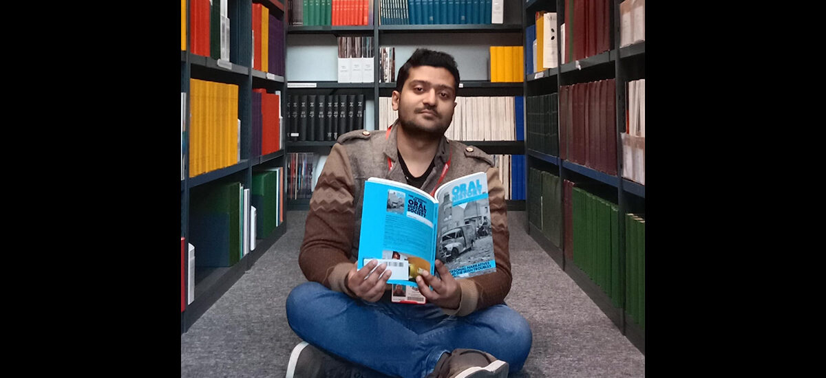 Student Sameer Iqbal sits cross-legged on the floor in a library aisle, holding and reading a magazine with colourful bookshelves in the background.