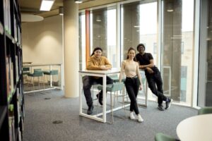 Three people are standing and sitting around a tall table in a modern, well-lit room with large windows and bookshelves.