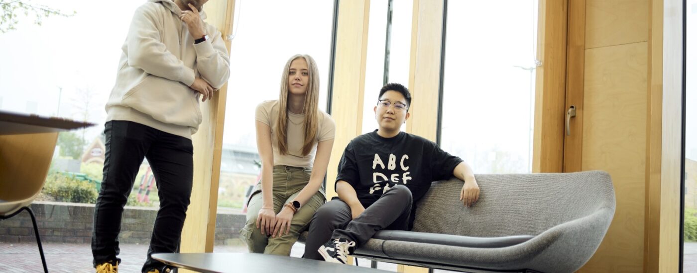 Three people pose in a modern, well-lit room with large windows; two are standing, and one is sitting on a grey sofa near a low table.