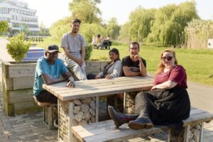 Five people sit and pose at a wooden outdoor picnic table in a park-like setting with trees and buildings visible in the background.