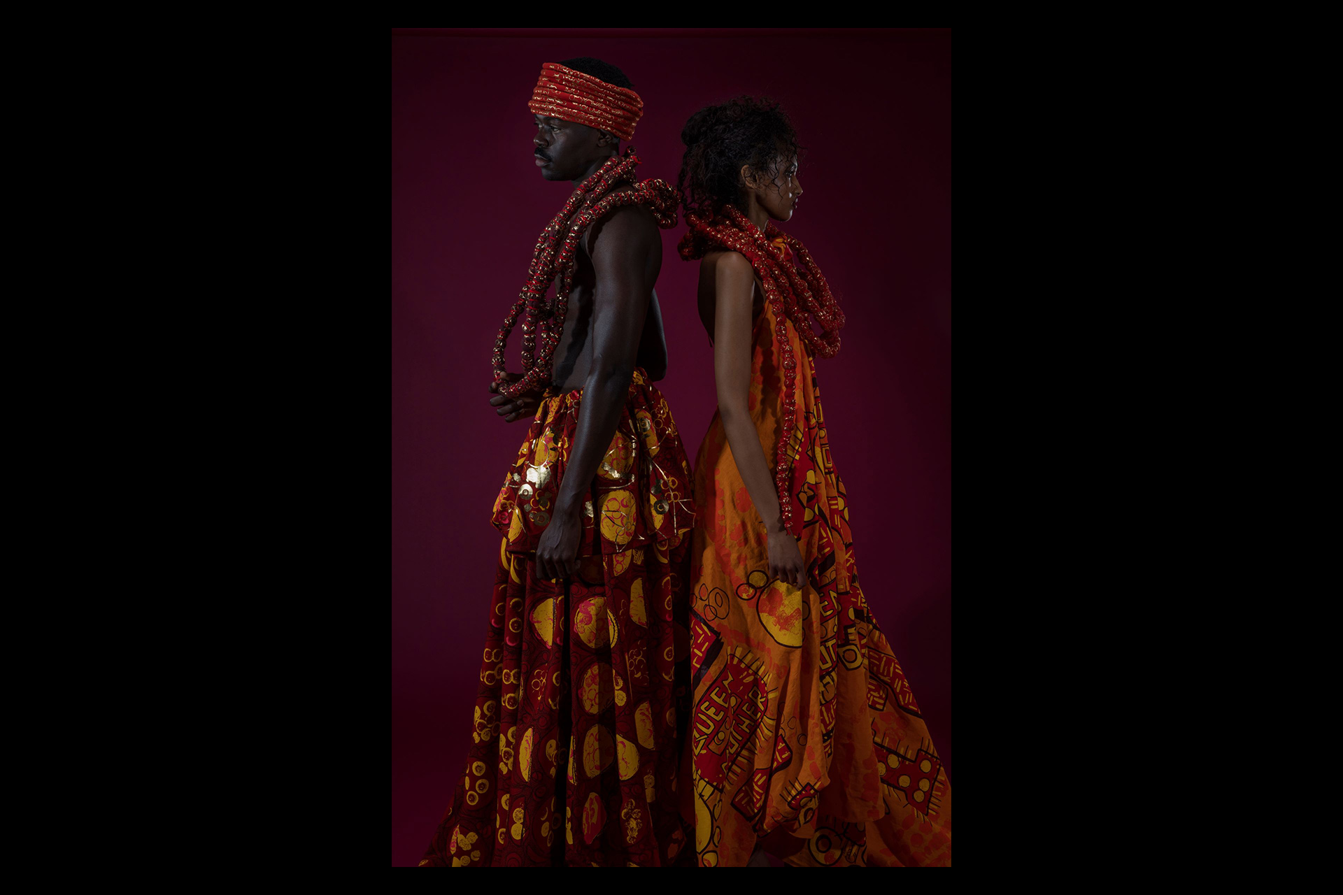 Two models stand back-to-back against a rich burgundy backdrop, wearing striking garments in vibrant red, orange, and yellow African-inspired prints. One model is shirtless, adorned with layered red beaded necklaces and a matching headwrap, while the other model wears a flowing dress paired with multiple strands of bold red beads.