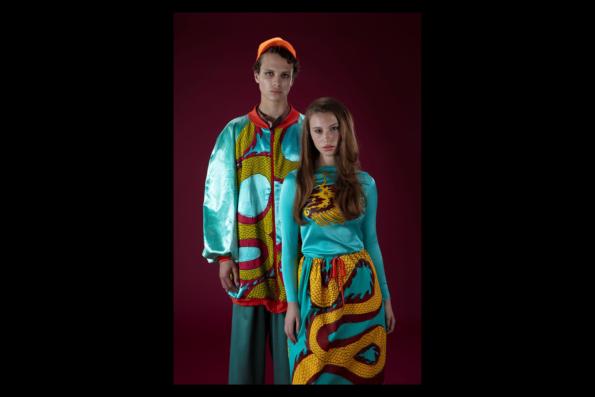 Two fashion models stand against a deep burgundy backdrop, wearing bold, colourful outfits featuring vibrant dragon motifs in turquoise, yellow, and red tones. One model wears a shiny bomber jacket and orange cap, while the other model dons a matching long-sleeve top and skirt with coordinated patterns.