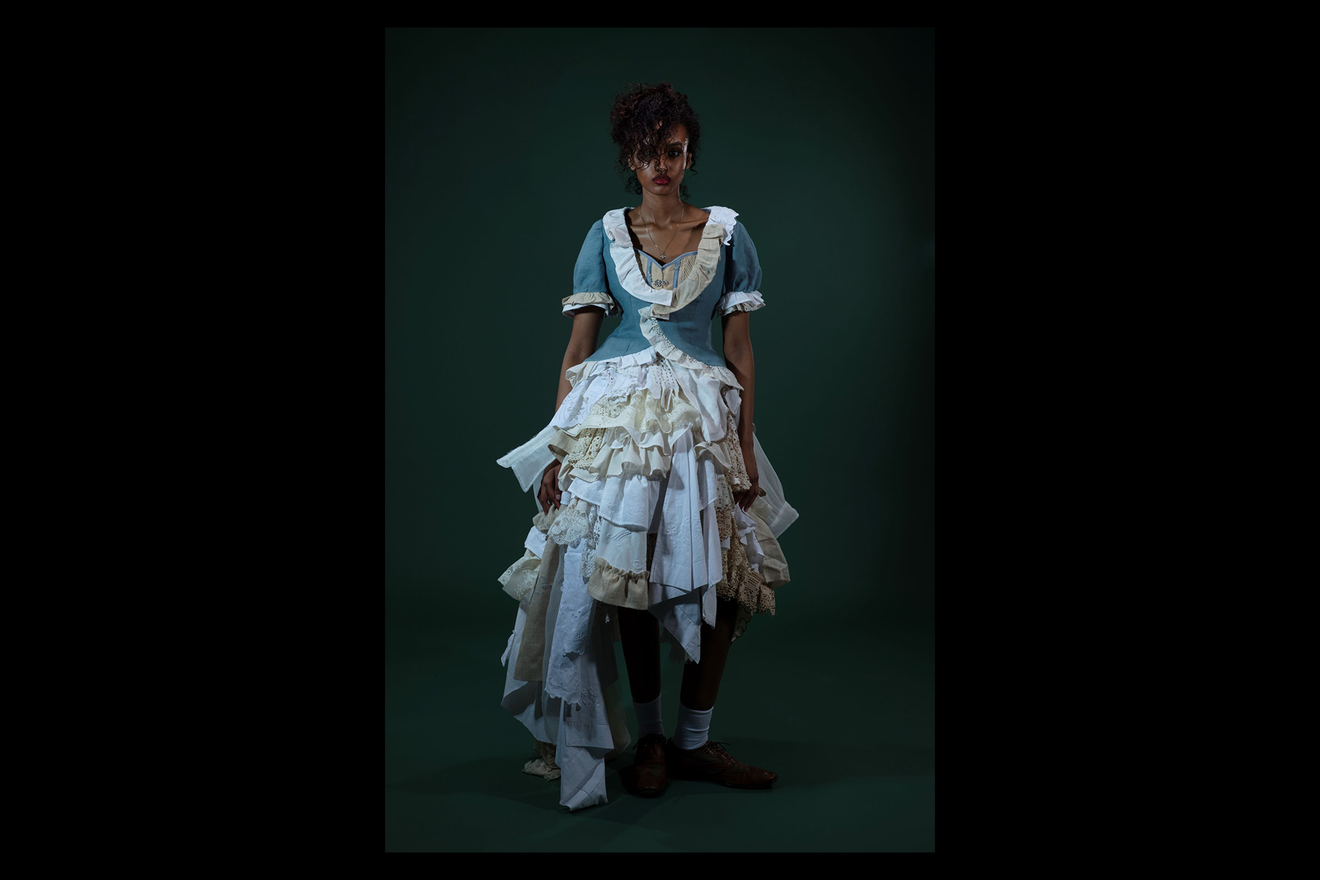 A model stands against a deep green backdrop wearing a theatrical, deconstructed historical costume. The outfit features a fitted blue bodice with cream ruffled trim and an elaborate, layered skirt made from various textured fabrics, lace, and linens.