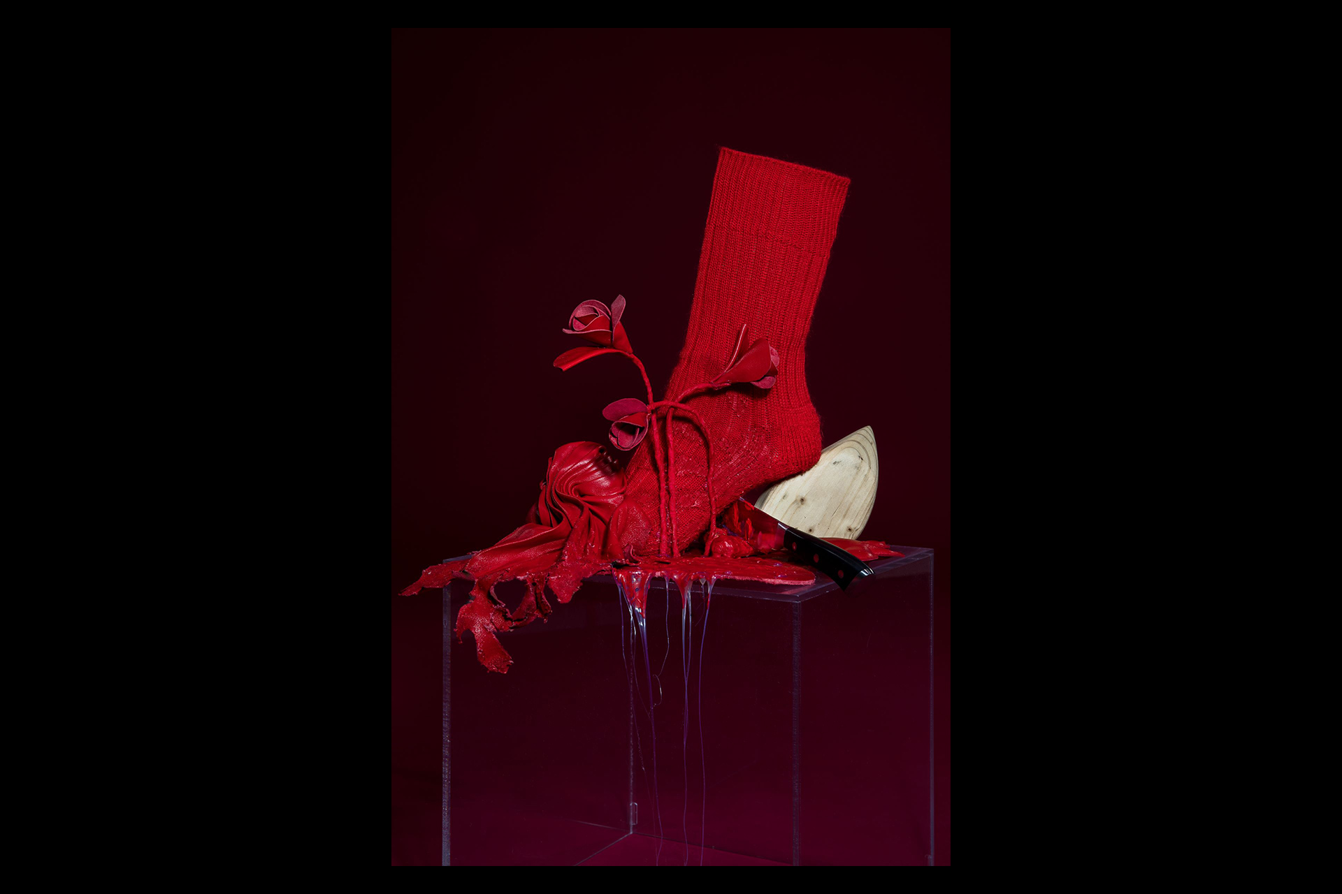 A conceptual fashion sculpture featuring a red knitted sock adorned with sculpted red roses, resting atop a melting red shoe form. The piece sits on a transparent plinth, with glossy red liquid dripping over the edges and a wooden shoe last and kitchen knife incorporated into the dramatic, surreal composition.
