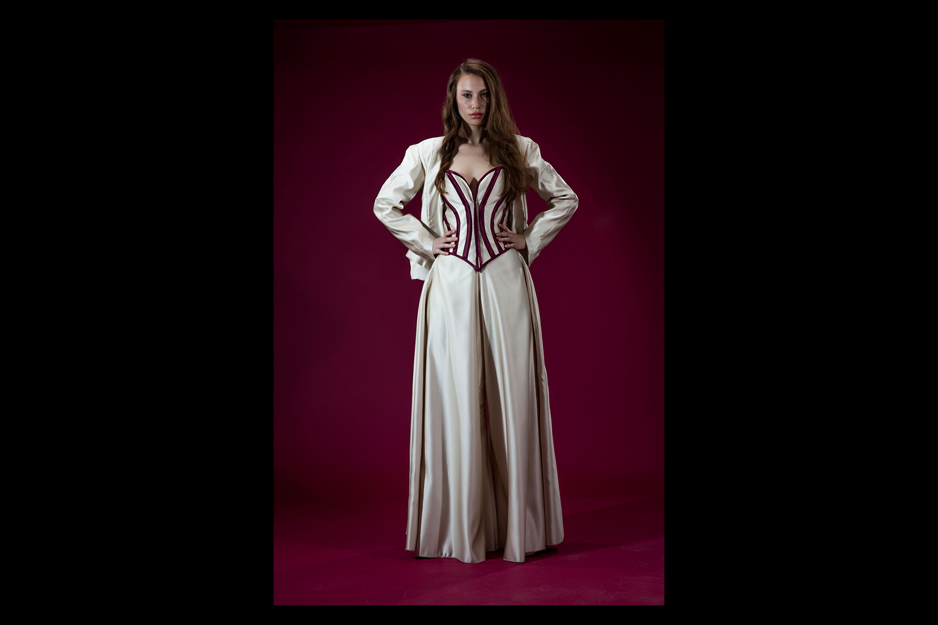 A model stands confidently against a rich burgundy background, wearing an elegant cream ensemble featuring wide-leg satin trousers, a structured corset with deep red velvet boning, and a matching cream jacket draped over the shoulders.