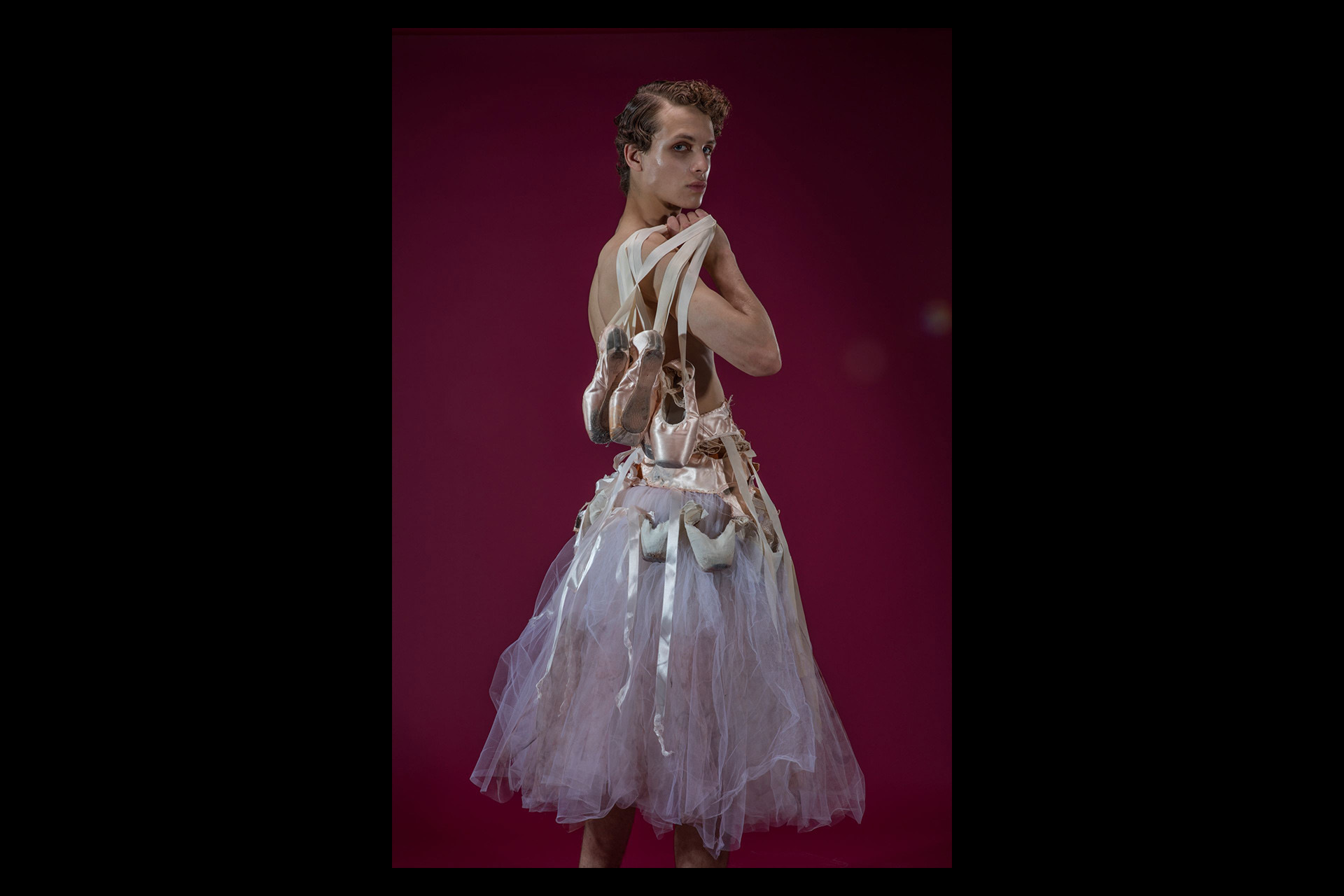 A model poses confidently against a deep burgundy background, wearing a dramatic tulle skirt adorned with worn ballet shoes and satin ribbons.