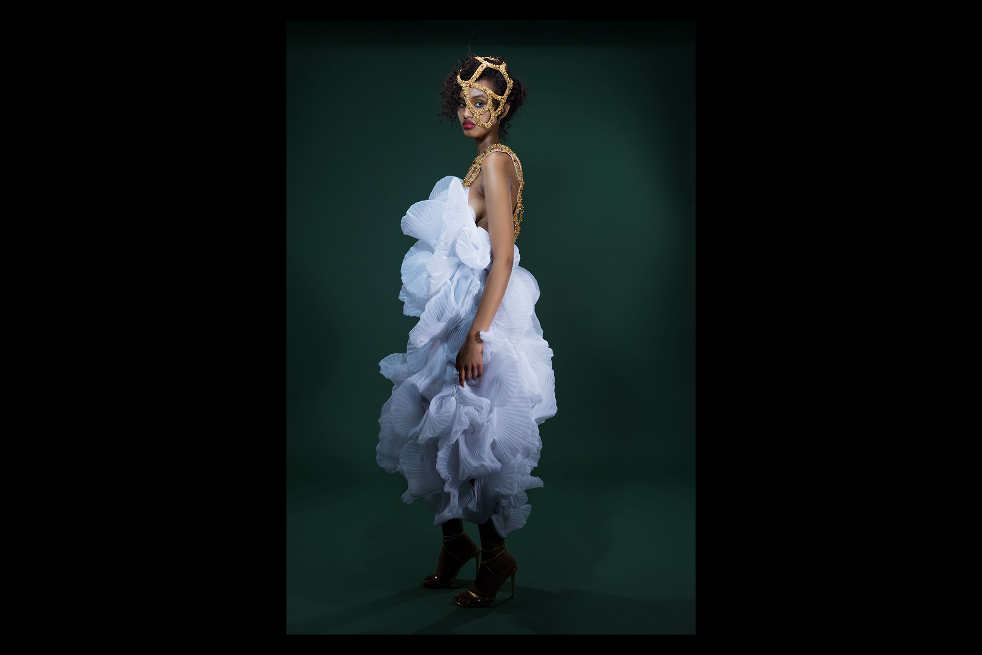 A model poses gracefully against a dark green backdrop, wearing a voluminous, sculptural white dress made of intricately pleated fabric. The look is accentuated by a striking gold embellished harness and matching ornate face mask.