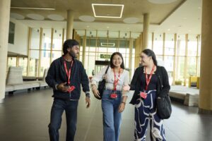 Three people wearing lanyards walk together and talk in a modern building lobby with large windows and geometric ceiling lights.