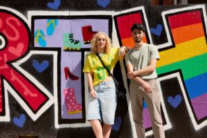 Two students stand in front of a colourful graffiti wall featuring boots, heels, and a rainbow section, posing casually for the camera.