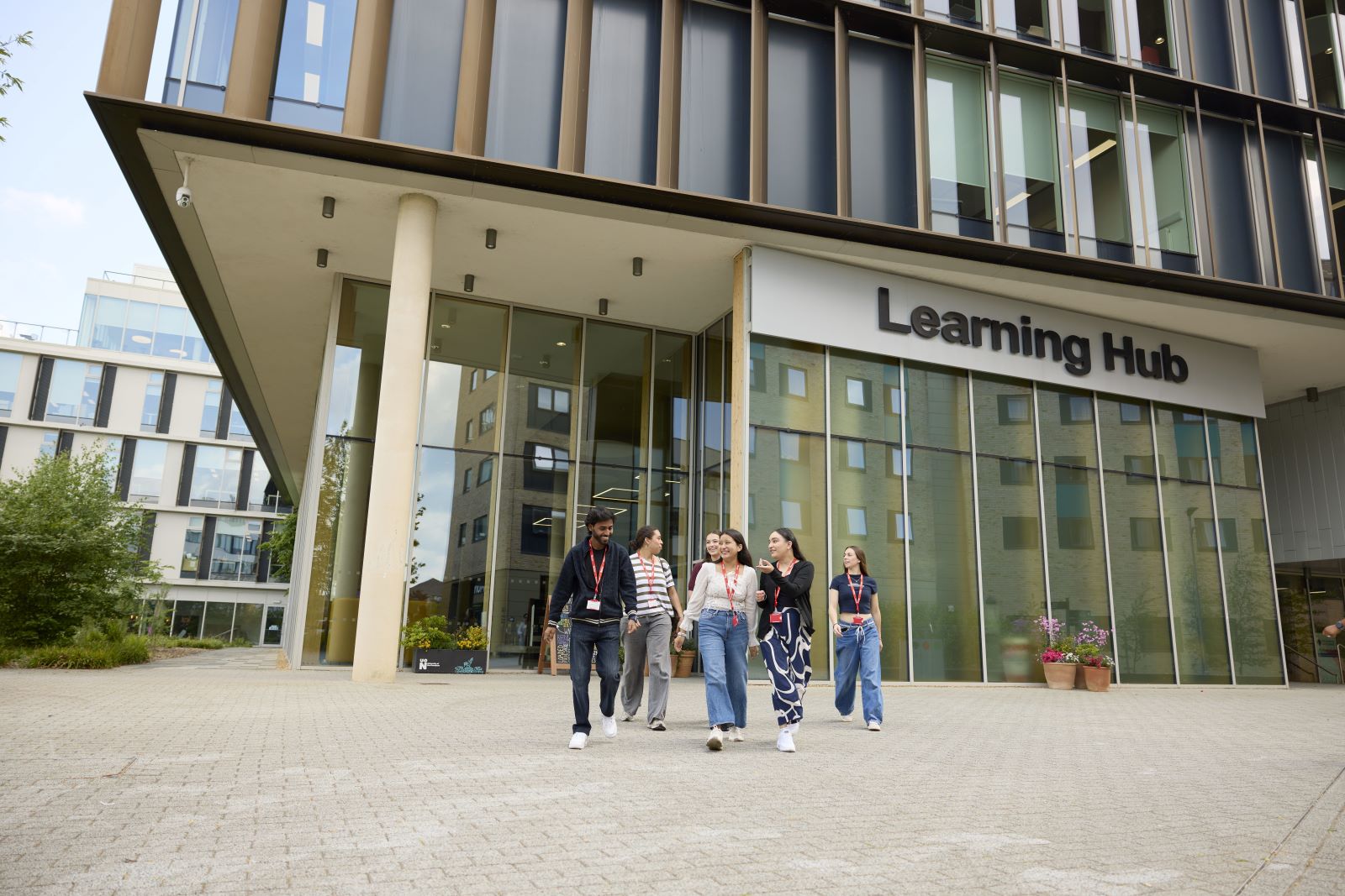 A group of students wearing red lanyards walk outside a modern building labeled "Learning Hub" with glass windows and pillars on UON's Waterside campus.