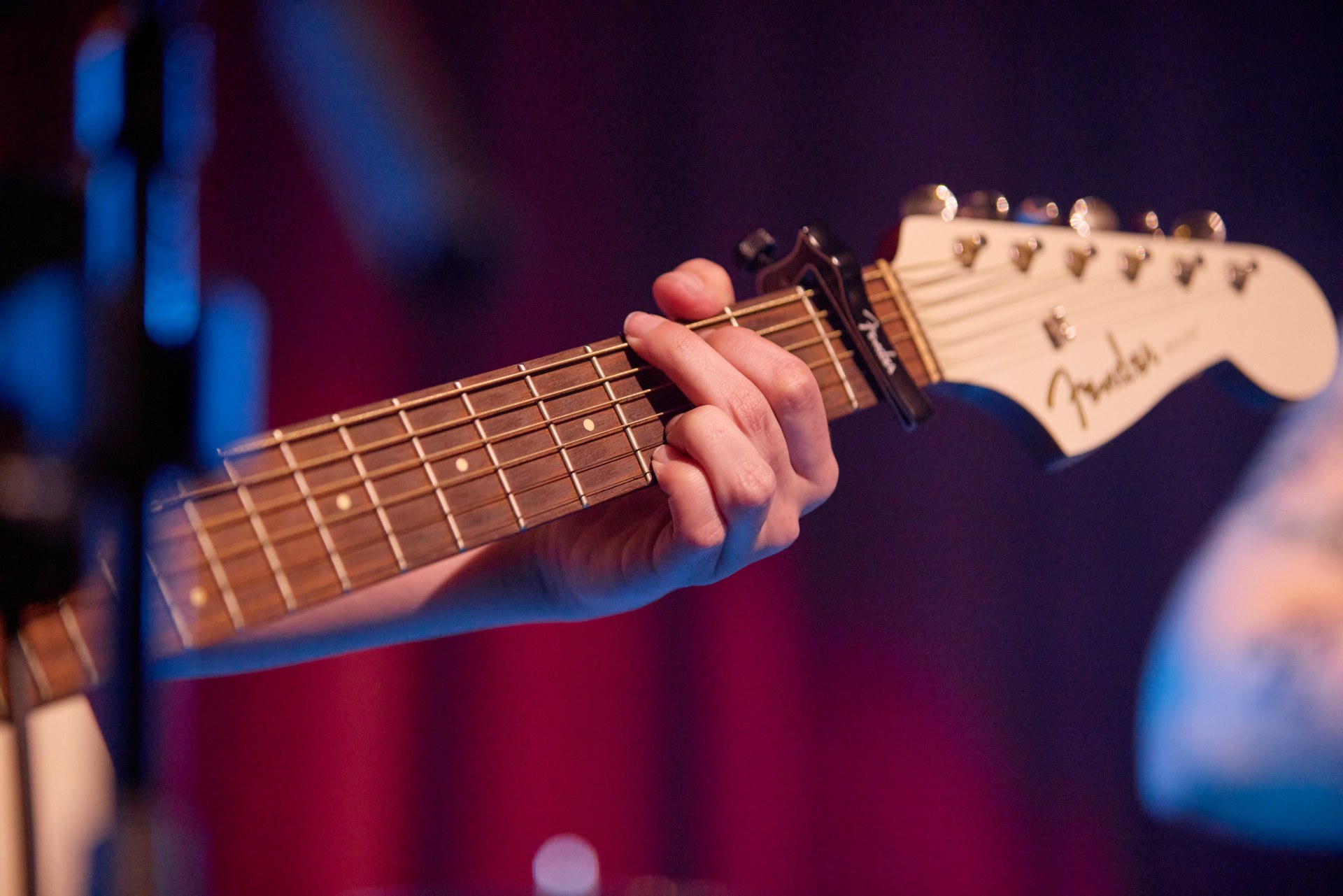 A close-up of a person's hand playing an electric guitar, with fingers pressing on the fretboard. The guitar headstock shows a Fender logo.