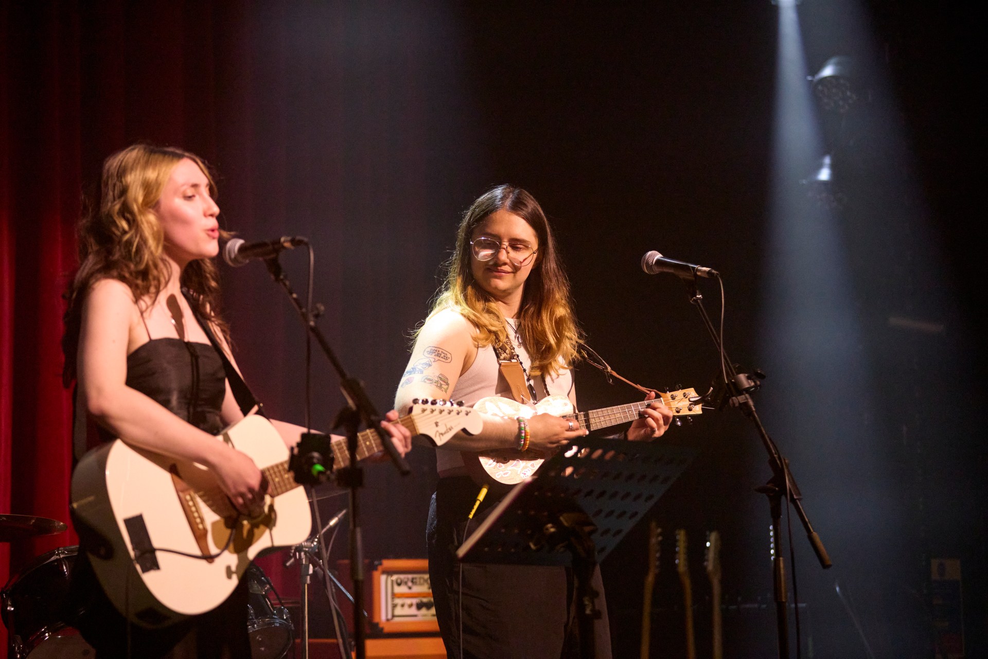 Two musicians perform on stage, one singing and playing an acoustic guitar, the other playing a ukulele. Red stage curtains and musical equipment are visible in the background.