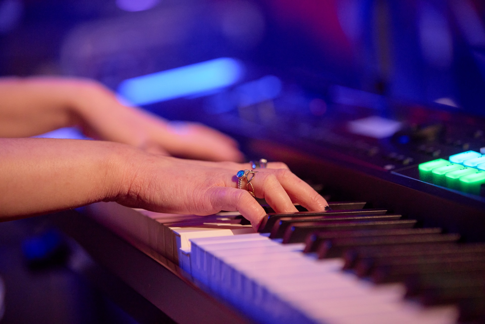 Close-up of a person's hands playing an electronic keyboard, with colored lighting and control buttons visible in the background.