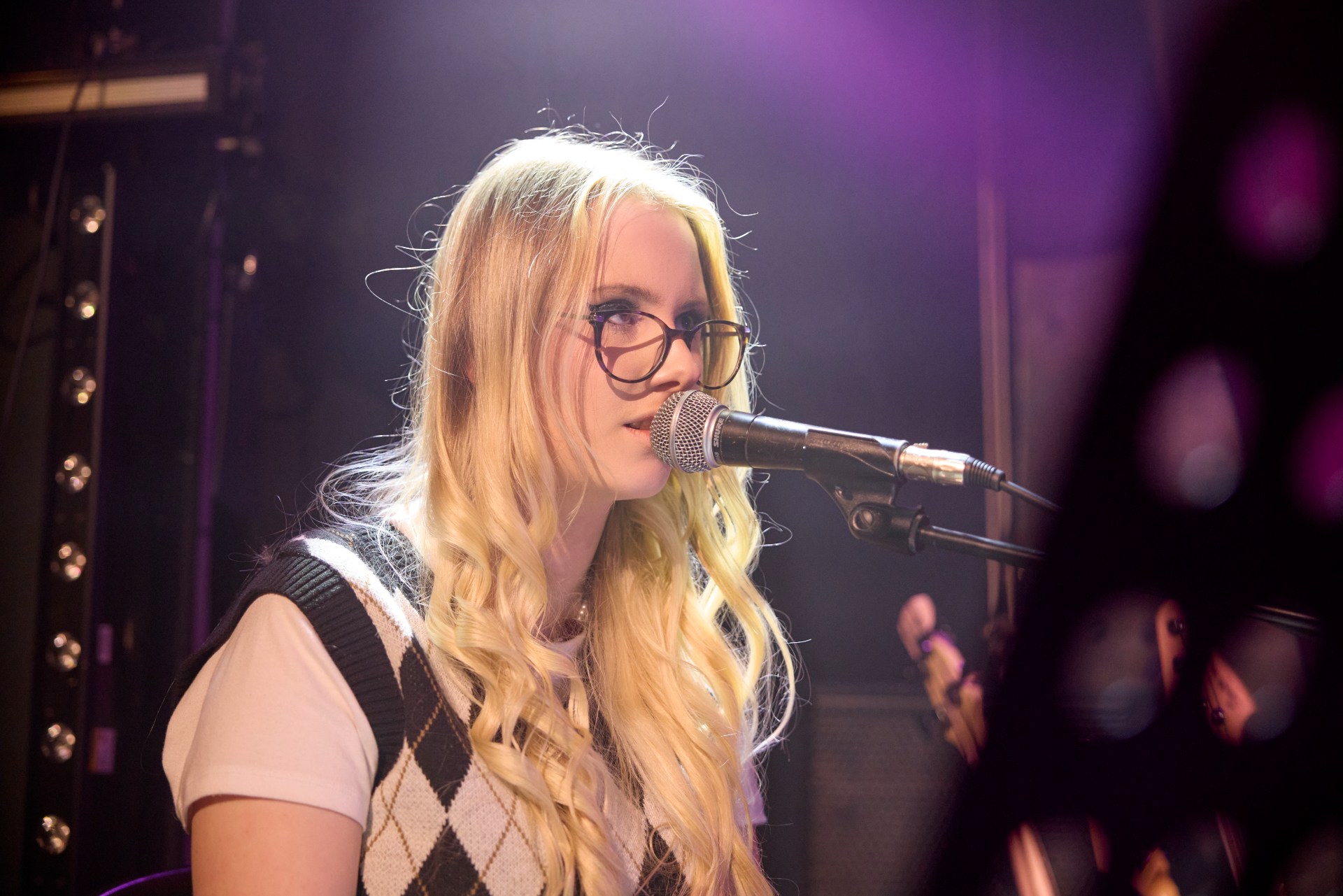 A blonde student with glasses sings into a microphone on stage, illuminated by purple lights.