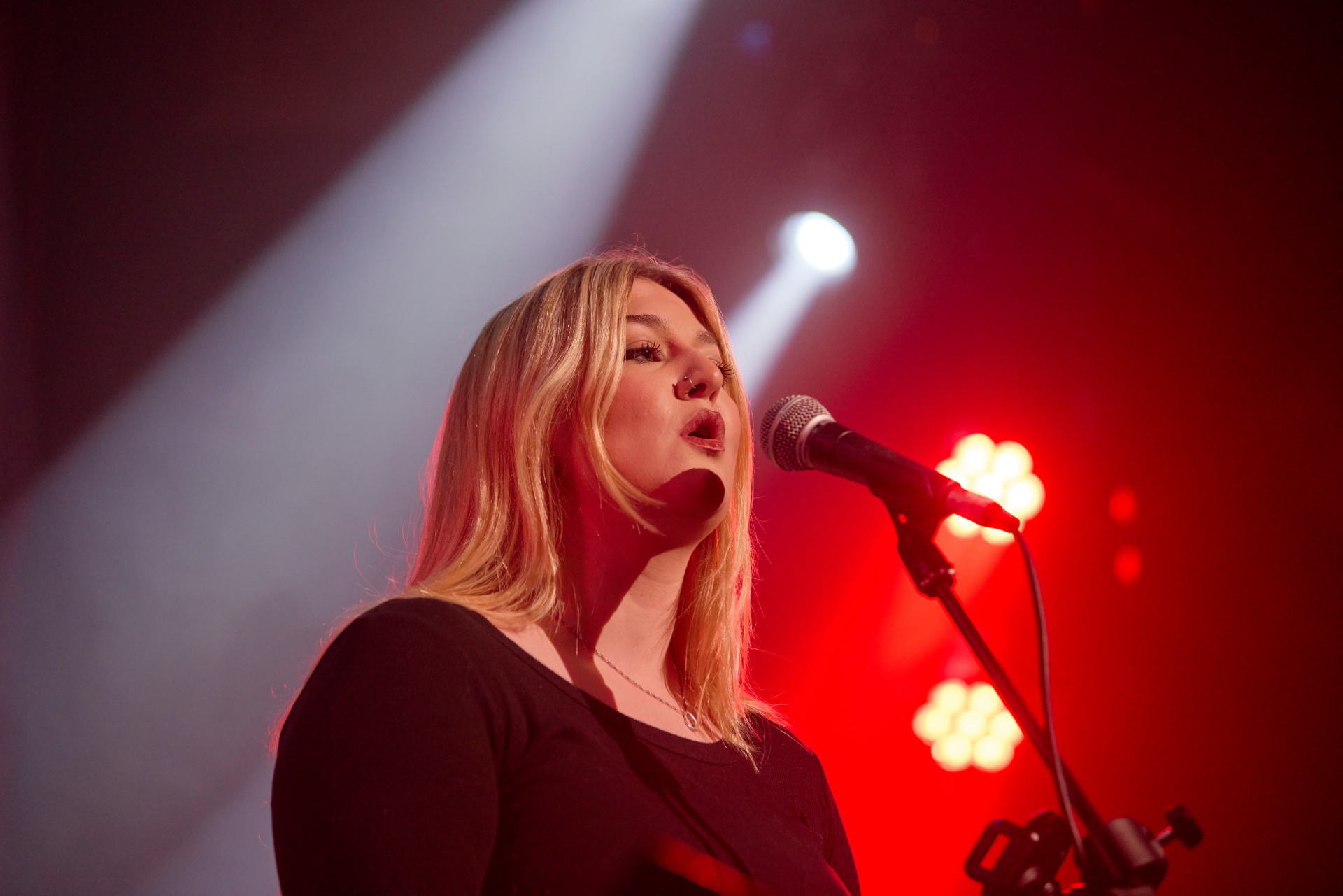 A woman with long blonde hair sings into a microphone on stage under red and white stage lights.