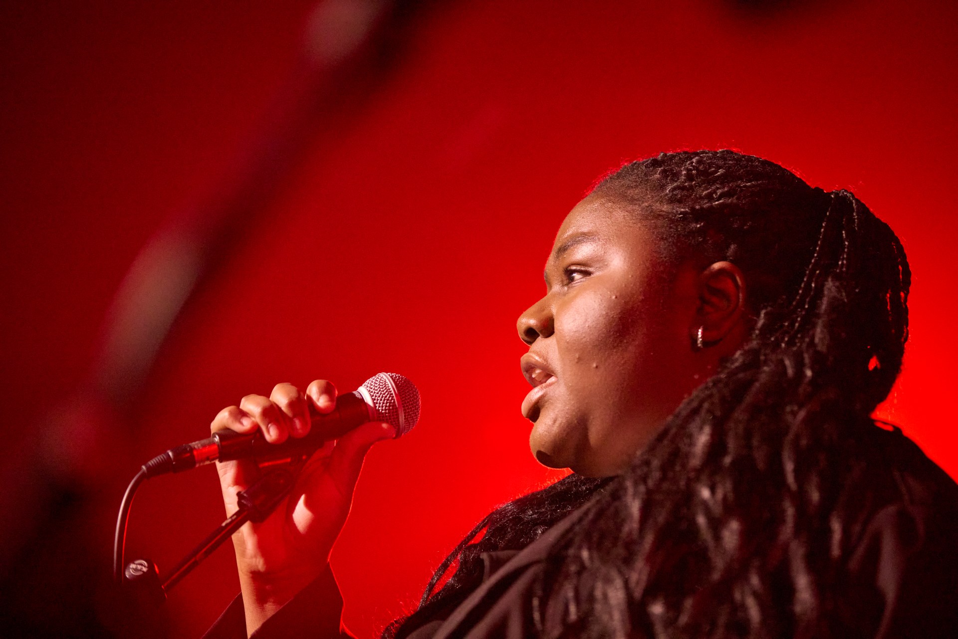 A person with long braids holds a microphone and sings on stage, illuminated by red lighting in the background.