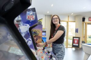 A young woman wearing glasses and a lanyard plays an arcade game in a brightly lit room with large windows.