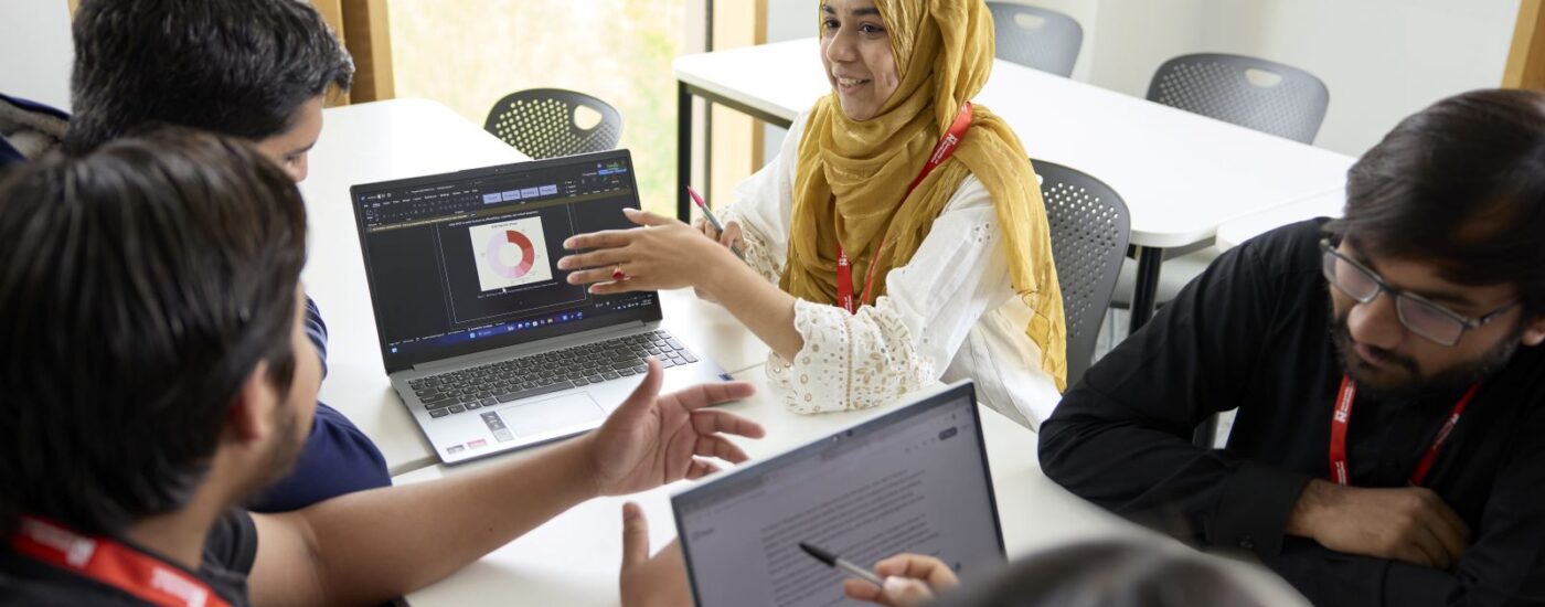 A group of students sit around a table in a classroom, discussing work on laptops, with one woman gesturing toward a screen displaying a diagram.