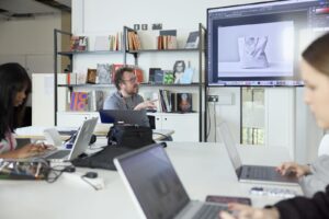 A group of people work on laptops at a table while a man presents a tote bag design on a large screen in a modern office setting.