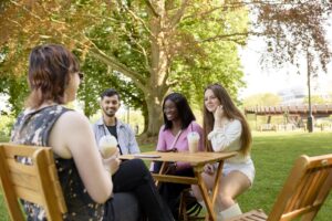 Four people sit at a wooden table outdoors in a park, talking and drinking beverages, with trees and sunlight in the background.