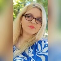 Image of Emilia Sarniak, wearing a blue and white patterned top, stands outdoors with greenery in the blurred background.