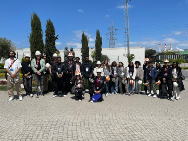 A group of people wearing safety helmets and visitor badges pose for a photo outdoors in front of industrial buildings and trees on a sunny day.