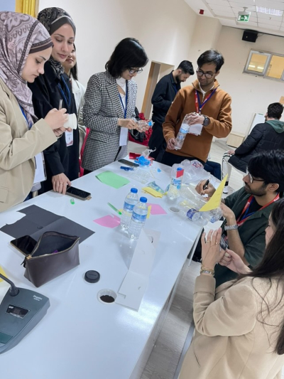A group of people are gathered around a table working on a craft project with paper, bottles, and other materials in a classroom setting.
