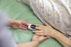 A nurse checks the oxygen level of a patient using a fingertip pulse oximeter while the patient lies in bed wearing a hospital property gown.