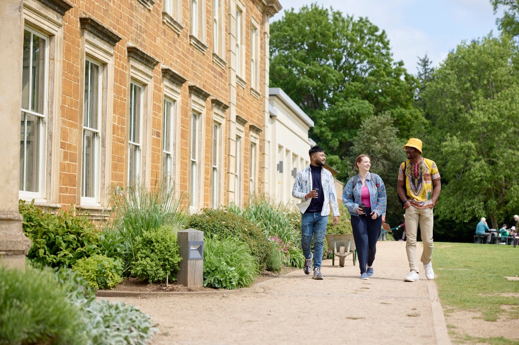 Three people walk and talk along a garden path beside a historic building, with green trees and additional people sitting in the background.