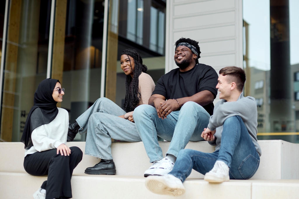 Four people sit on outdoor steps, talking and laughing together in front of a modern building.