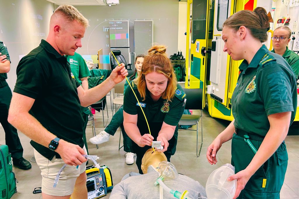 Three paramedics practice intubation on a medical mannequin in a training room, with medical equipment and an ambulance visible in the background.
