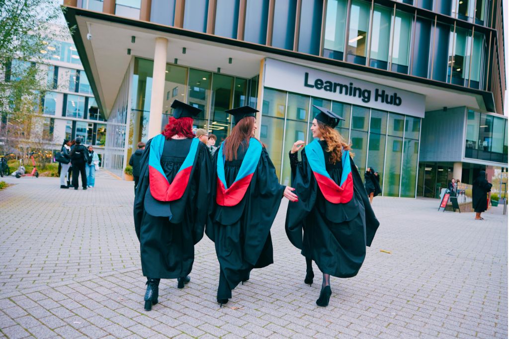 Three graduates in caps and gowns walk toward a building labeled "Learning Hub," with other people visible in the background.