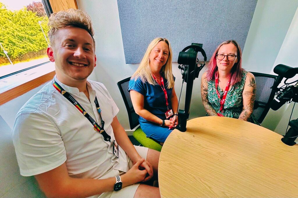Three people sit around a round table with microphones in a brightly lit room, suggesting they are recording a podcast or interview.