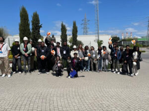 A group of people, some wearing hard hats and holding safety helmets, pose for a photo outdoors on a paved area with trees and industrial buildings in the background.