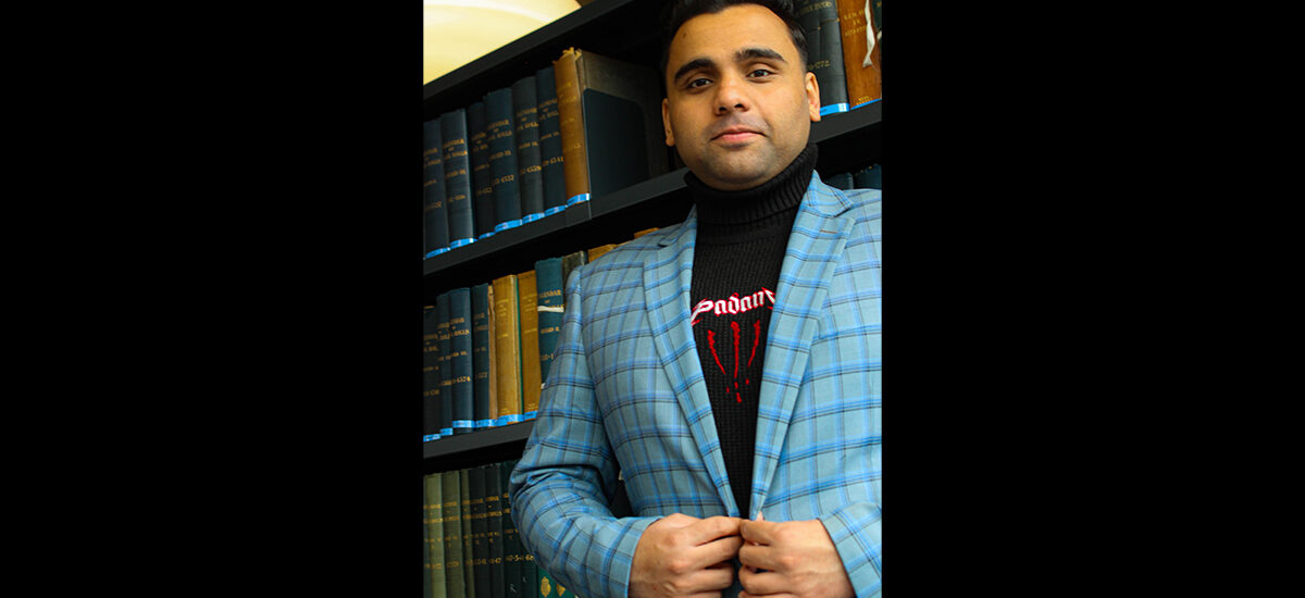 Muhammad Ayan Rao, Project Management MSc graduate, wears a blue plaid blazer and black turtleneck stands in front of a bookshelf filled with old books.