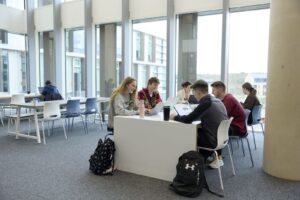 Students sit around a table studying with laptops and notebooks in a modern, light-filled campus common area with large windows.
