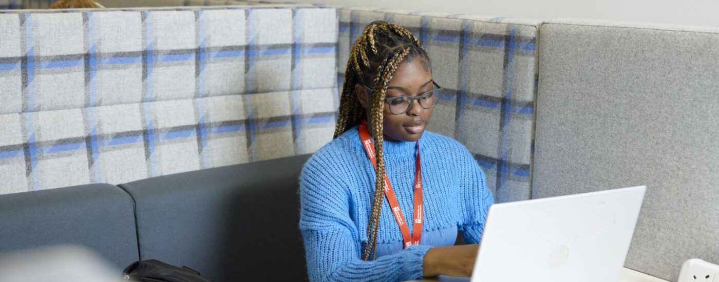 Decorative image of A woman wearing glasses and a blue sweater works on a laptop at a table in a booth with grey and blue padded walls.