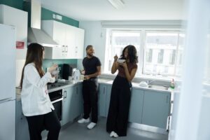 Three people stand and eat from bowls in a modern kitchen, with appliances, cupboards, and large windows visible in the background.