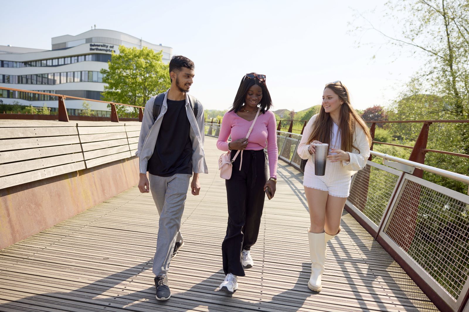 Three young students walk together on a wooden bridge in daylight, chatting and holding drinks, with UON's Creative Hub and trees in the background.